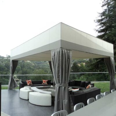 a beautiful patio with three black sofas and two white chairs covered in a white patio awning with gray curtains with green scenery - patio awning options - patio awnings - patio outdoor awnings - patio shades - custom awnings - custom shape awnings near me