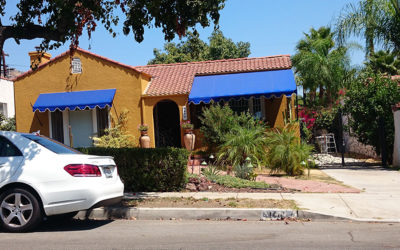 custom blue awnings in a yellow one-story house with plants in the front yard and a parked white car - standard awnings - concave awnings - patio shades - custom awnings - custom awnings for patio - custom awnings covers