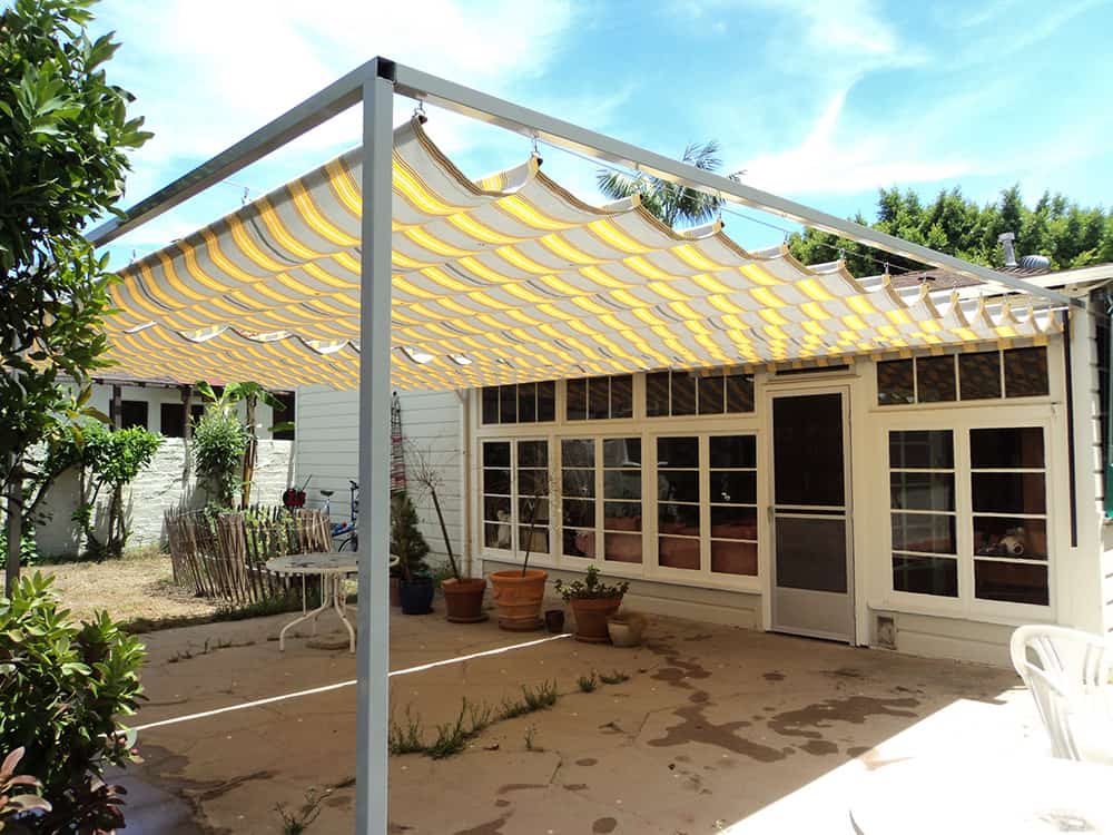 patio with stripe yellow slide on wire retractable awning with plants and a table - custom awnings - custom aluminum awnings - custom awning designs - custom awnings cost - retractable awnings - spear awnings - convex awnings - patio shades