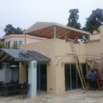 a man with a ladder installing awning on a yellow house - custom awnings - custom awnings near me - custom awnings for decks - custom awnings for business - custom door awnings near me - spear awnings - aluminum awnings - dome awnings