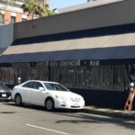 two men installing storefront awning for a restaurant with two cars parked in front - custom awnings - custom awnings near me - custom awnings for decks - custom awnings for business - custom door awnings near me - spear awnings - aluminum awnings - dome awnings
