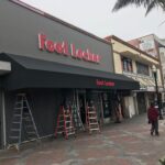 two men installing a black awning for a foot locker store - custom awnings - custom awnings near me - custom awnings for decks - custom awnings for business - custom door awnings near me - spear awnings - aluminum awnings - dome awnings