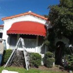 a white house with a red dome awning and a man in front with a ladder - custom awnings - custom awnings near me - custom awnings for decks - custom awnings for business - custom door awnings near me - spear awnings - aluminum awnings - dome awnings