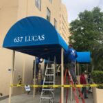two men installing a blue canopy for the building - custom awnings - custom awnings near me - custom awnings for decks - custom awnings for business - custom door awnings near me - spear awnings - aluminum awnings - dome awnings