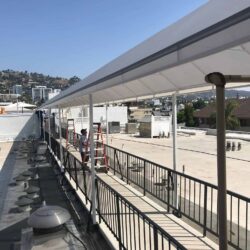 two men installing grey entrance canopy for a white building with flags in the background - custom awnings - custom awnings near me - custom awnings for decks - custom awnings for business - custom door awnings near me - spear awnings - aluminum awnings - dome awnings