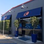 blue storefront awning for a grey store with plants on the entrance - custom awnings - custom awnings near me - custom awnings for decks - custom awnings for business - custom door awnings near me - spear awnings - aluminum awnings - dome awnings
