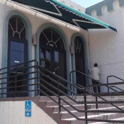 cyan retractable storefront awnings for a white and light blue store with a front stairway and a man wearing a white shirt - custom store front awnings - retractable awnings - restaurant awnings - standard awnings - patio enclosures - commercial awnings - storefront awning cost - storefront with awning