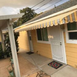 stripe yellow straight awnings for a yellow house with a white entrance door - custom awnings - custom awnings near me - custom awnings for decks - custom awnings for business - custom door awnings near me - spear awnings - aluminum awnings - dome awnings