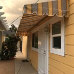 stripe yellow straight awnings for a yellow house with a white entrance door - custom awnings - custom awnings near me - custom awnings for decks - custom awnings for business - custom door awnings near me - spear awnings - aluminum awnings - dome awnings