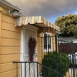 stripe yellow straight awnings for a yellow house with a white entrance door with a christmas decoration- custom awnings - custom awnings near me - custom awnings for decks - custom awnings for business - custom door awnings near me - spear awnings - aluminum awnings - dome awnings