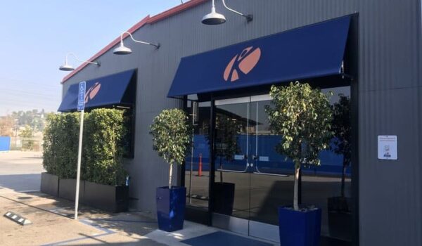 blue storefront awning for a grey store with plants on the entrance - custom awnings - custom awnings near me - custom awnings for decks - custom awnings for business - custom door awnings near me - spear awnings - aluminum awnings - dome awnings