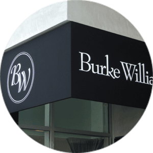 A black awning above a building entrance displays “Burke Williams” in white letters on the right and a circular “BW” logo on the left. The awning is attached to a modern building with glass windows beneath it.