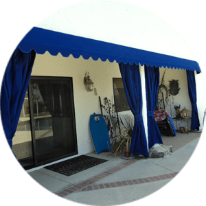 A patio area with a white wall, sliding glass door, and window features a deep blue fabric awning and matching blue curtains tied to columns. Various outdoor items and plants are placed near the wall, and a swimming pool is visible through the glass door.