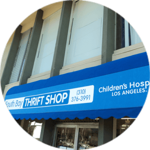 A blue awning above a storefront reads “South Bay THRIFT SHOP” with a phone number in the center, and “Children’s Hospital Los Angeles” on the right. The building behind has large windows and beige columns.