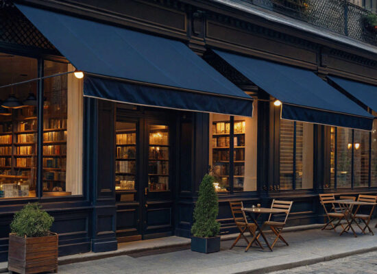A cozy café with custom awnings in dark blue and large windows warmly lit from the inside. Wooden tables and chairs rest on the stone sidewalk, flanked by potted plants. The street and building exterior have a classic, European style.