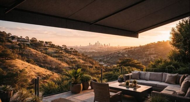 A modern patio with a large sectional sofa, chairs, and potted plants overlooks a sunlit, hilly landscape at sunset. A city skyline is visible in the distance beneath a dramatic, extended awning. The scene feels relaxed and luxurious.